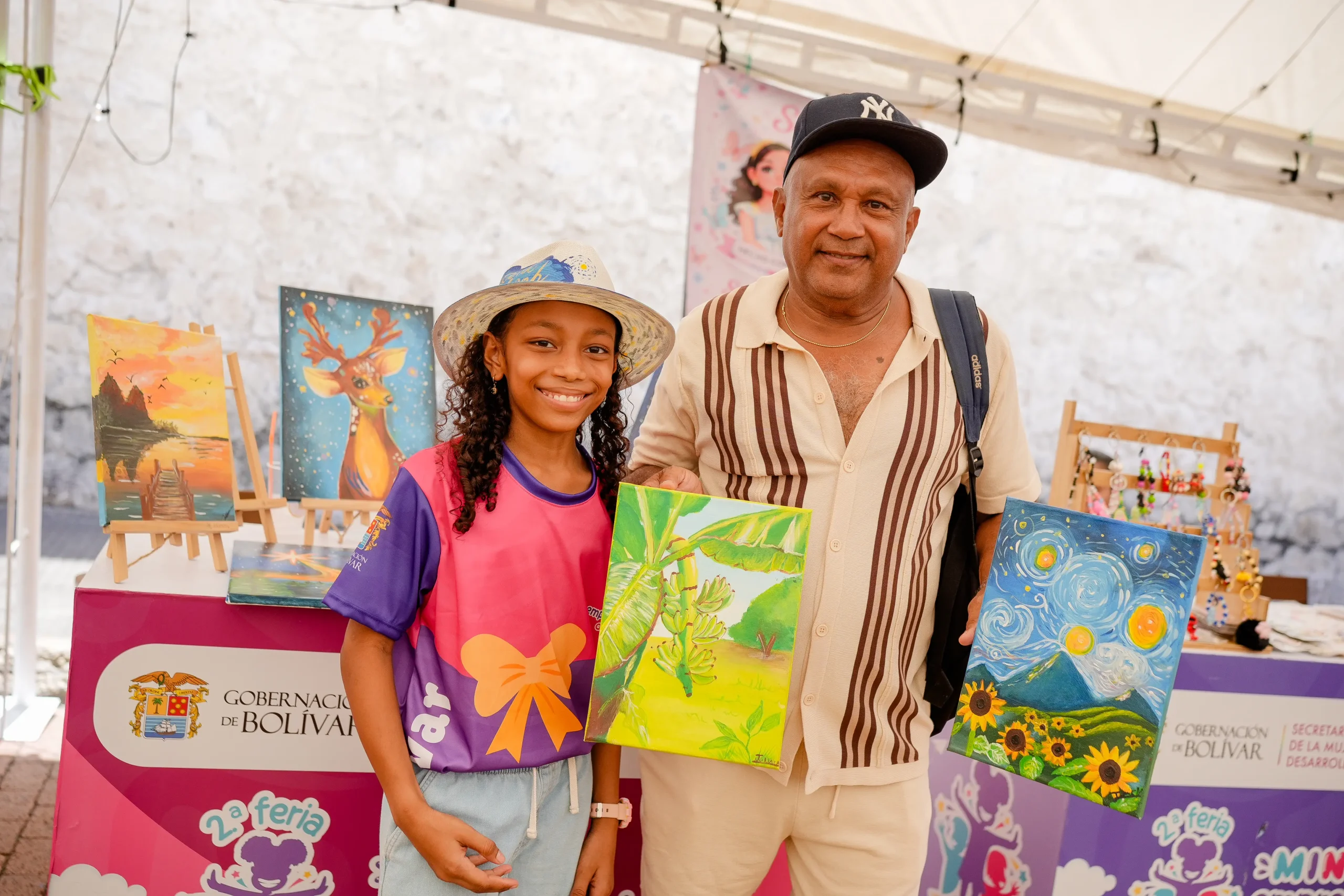 Niña exhibe pinturas junto a un adulto acompañante