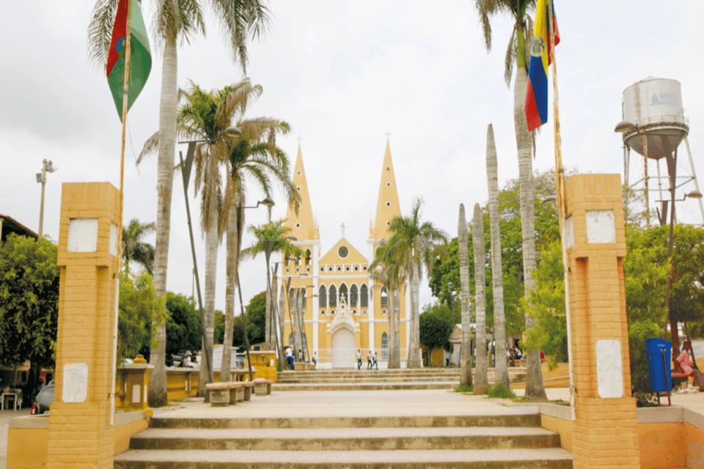Fotografía panorámica de la plaza principal de Turbaco, Bolívar. En primer plano se observan senderos peatonales, palmeras y astas con banderas, mientras que al fondo destaca la iglesia principal del municipio con su característica fachada de tonos claros y torres puntiagudas. La imagen refleja el patrimonio arquitectónico, cultural y turístico de la población.