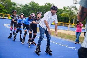 Patinadores con casco y uniforme recorren la renovada pista de patinaje azul junto a un hombre de camisa blanca.