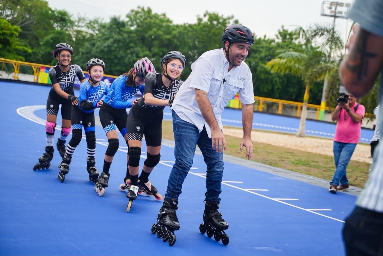 Patinadores con casco y uniforme recorren la renovada pista de patinaje azul junto a un hombre de camisa blanca.