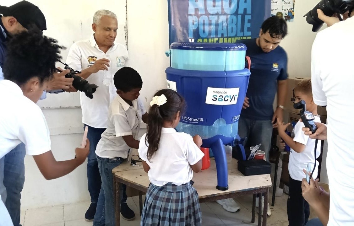 Niños y niñas con uniforme escolar junto a un filtro purificador de agua azul, mientras adultos registran el momento con cámaras.