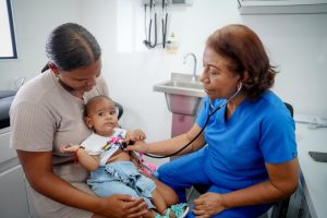 Enfermera con estetoscopio examina a un bebé en brazos de su madre dentro de un consultorio.