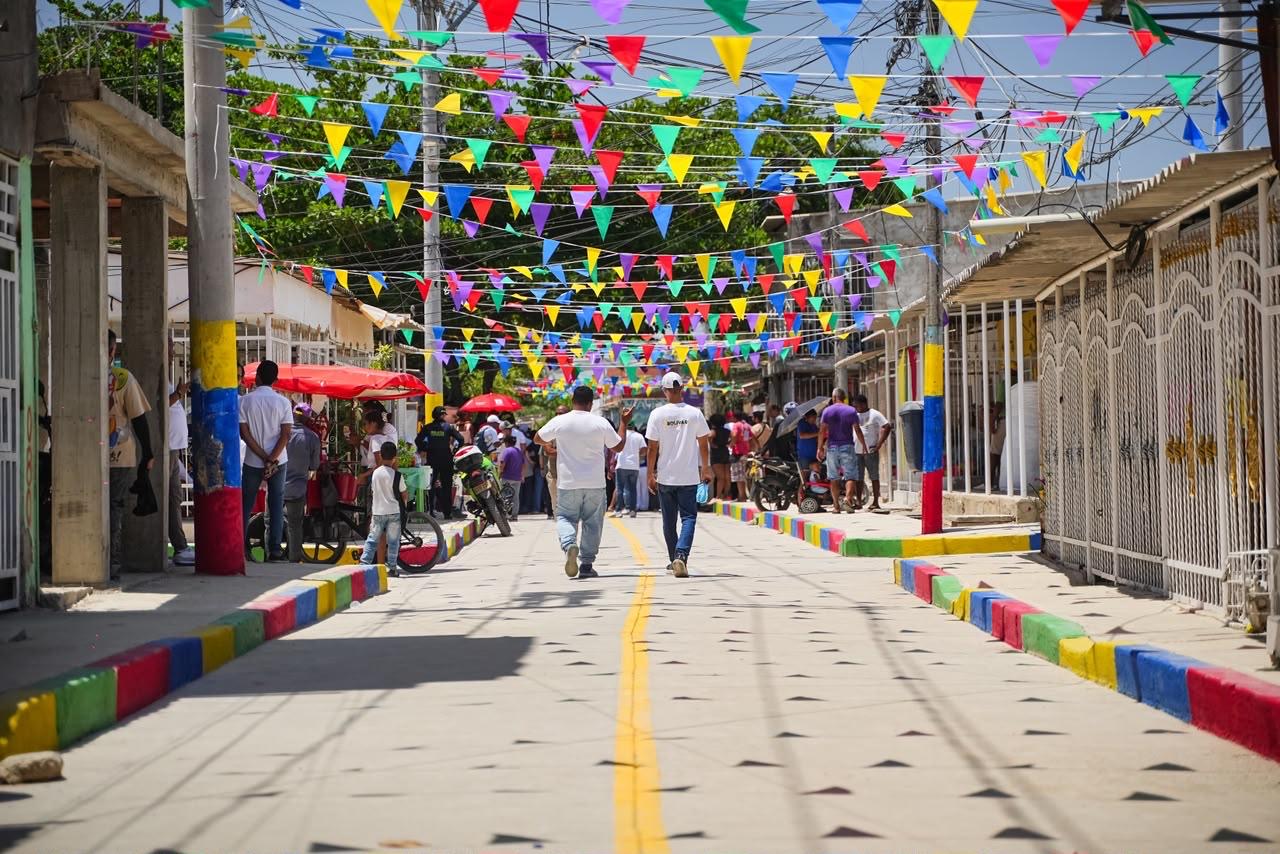 Calle recién pavimentada con bordillos pintados de colores y banderines festivos, con personas caminando.