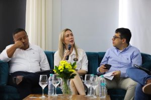 Tres personas sentadas en un sofá durante un conversatorio; una mujer habla con micrófono junto a una mesa con flores.