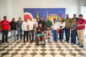 Grupo de personas posa frente a un telón con el escudo de Bolívar y banderas, en un salón de piso ajedrezado; una de ellas en silla de ruedas.
