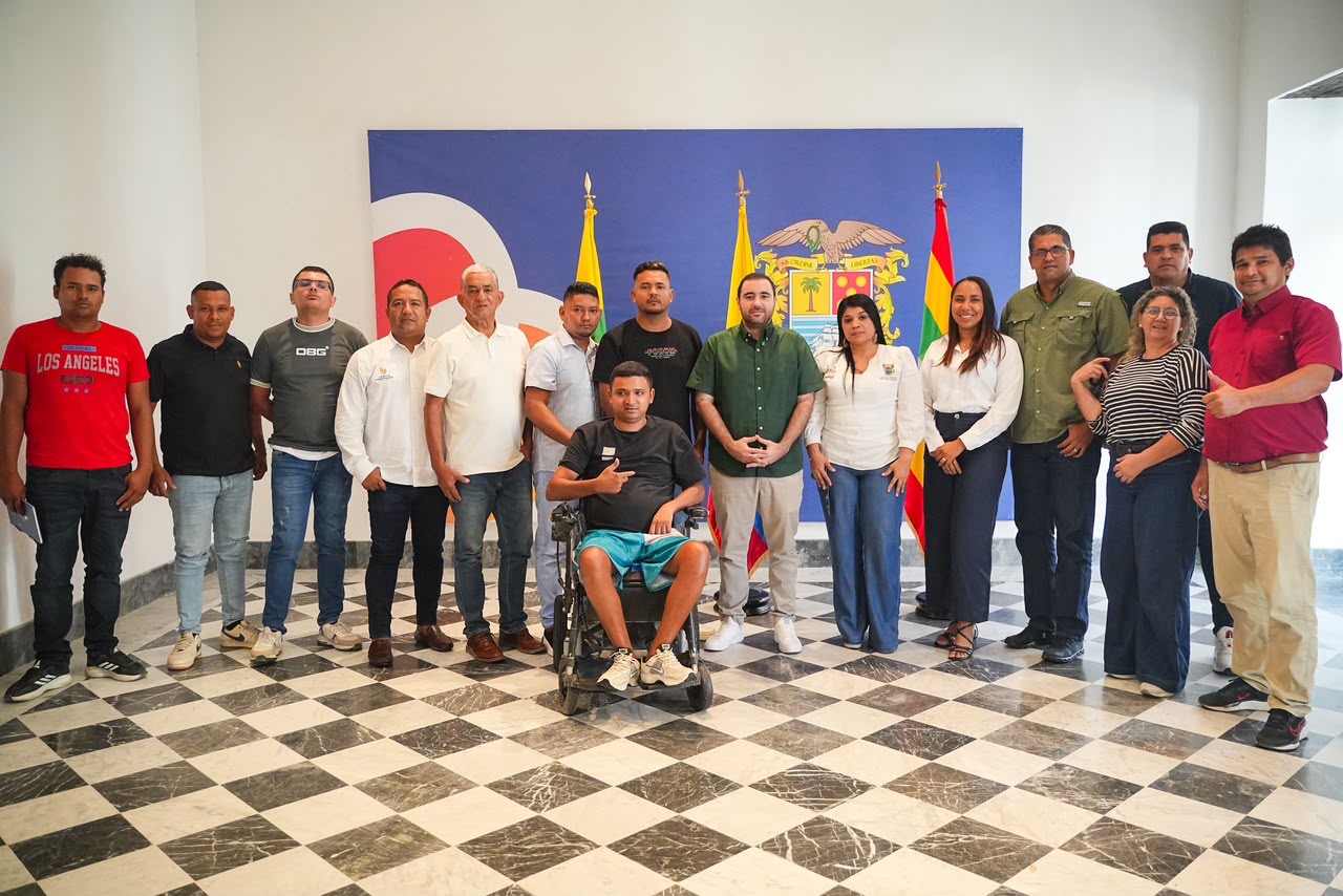 Grupo de personas posa frente a un telón con el escudo de Bolívar y banderas, en un salón de piso ajedrezado; una de ellas en silla de ruedas.