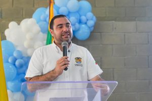 Hombre de camisa blanca habla con micrófono frente a una decoración de globos azules y blancos.