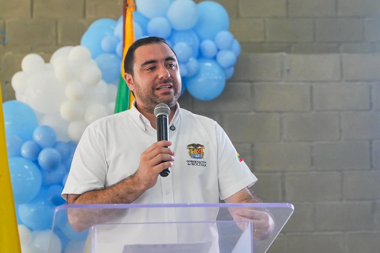 Hombre de camisa blanca habla con micrófono frente a una decoración de globos azules y blancos.