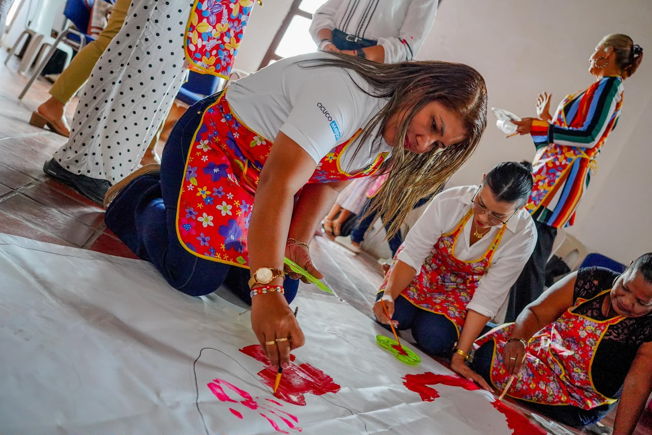 Mujeres con delantales de colores pintan con las manos un mural sobre papel en el piso.