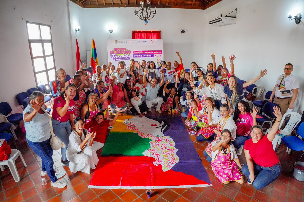 Grupo de mujeres celebra con los brazos en alto alrededor de un mural pintado en el piso de un salón.