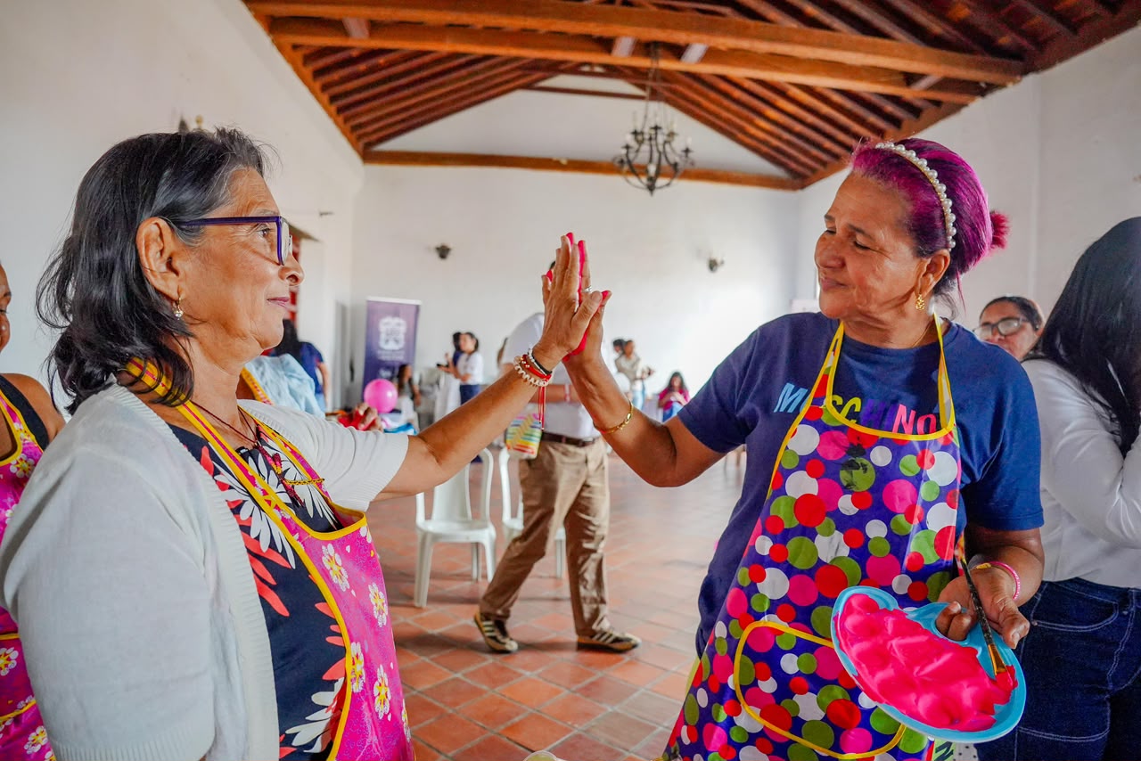 Dos mujeres mayores con delantales chocan las manos durante una actividad en un salón.