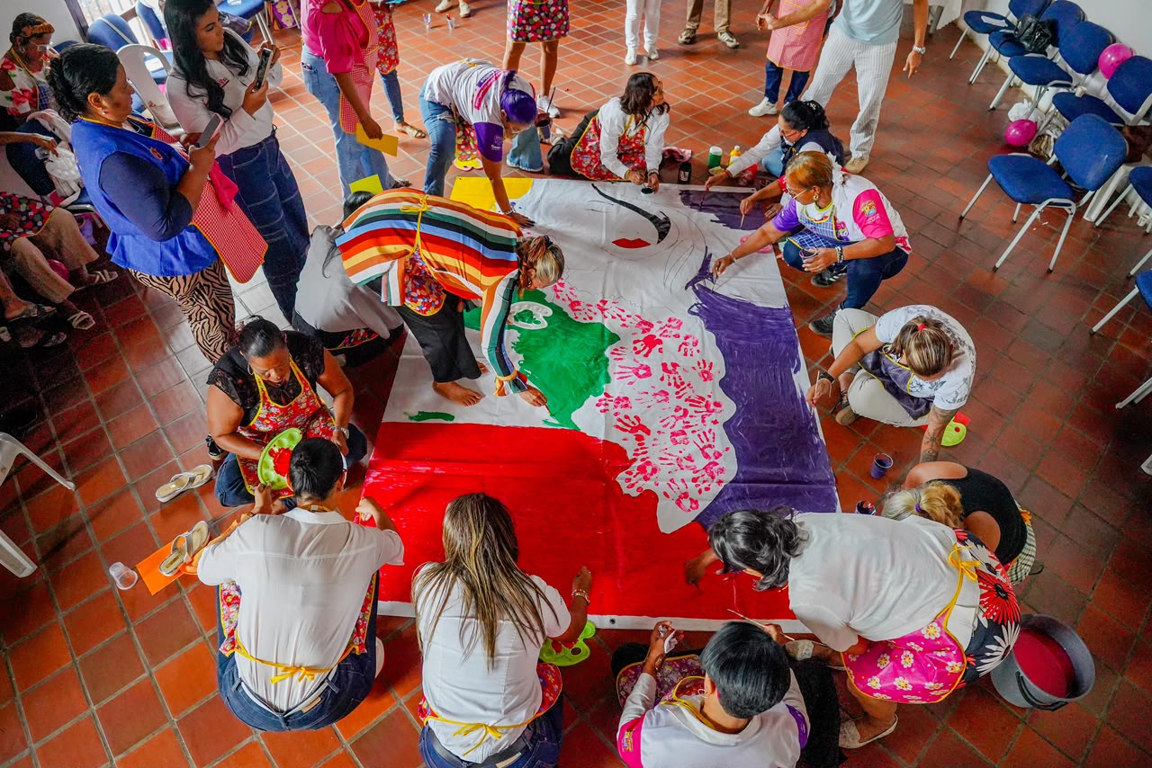 Vista desde arriba de mujeres que pintan un gran mural colorido sobre el piso del salón.