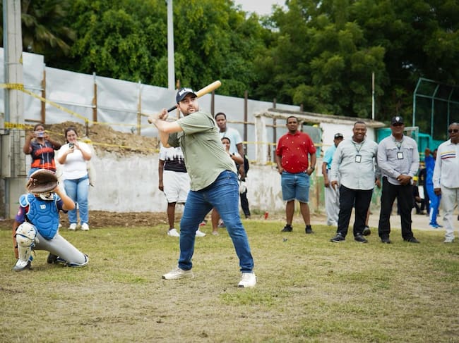 Hombre batea en un campo de béisbol en obra mientras un grupo de personas observa.