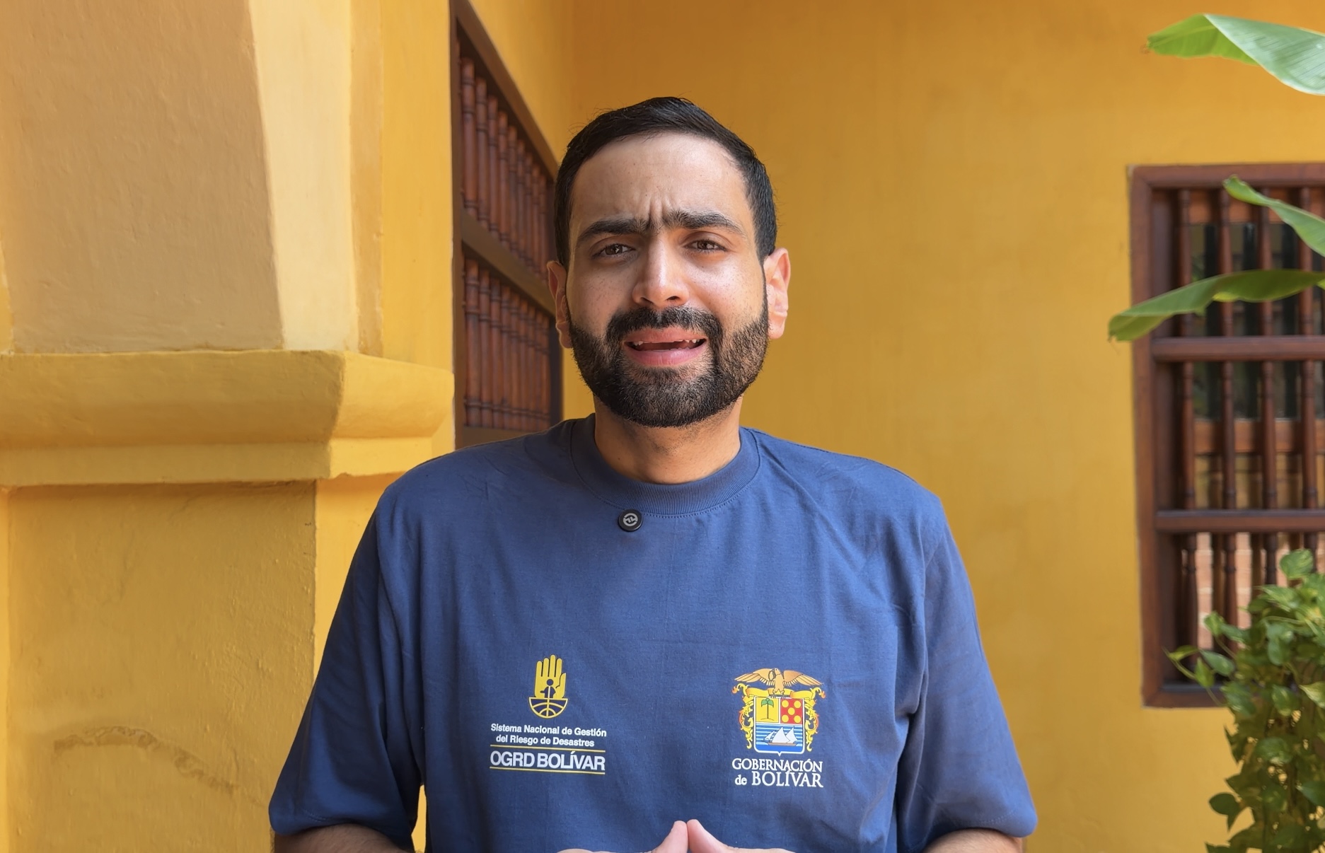 Hombre con camiseta azul de la Gobernación de Bolívar habla frente a una fachada colonial amarilla.