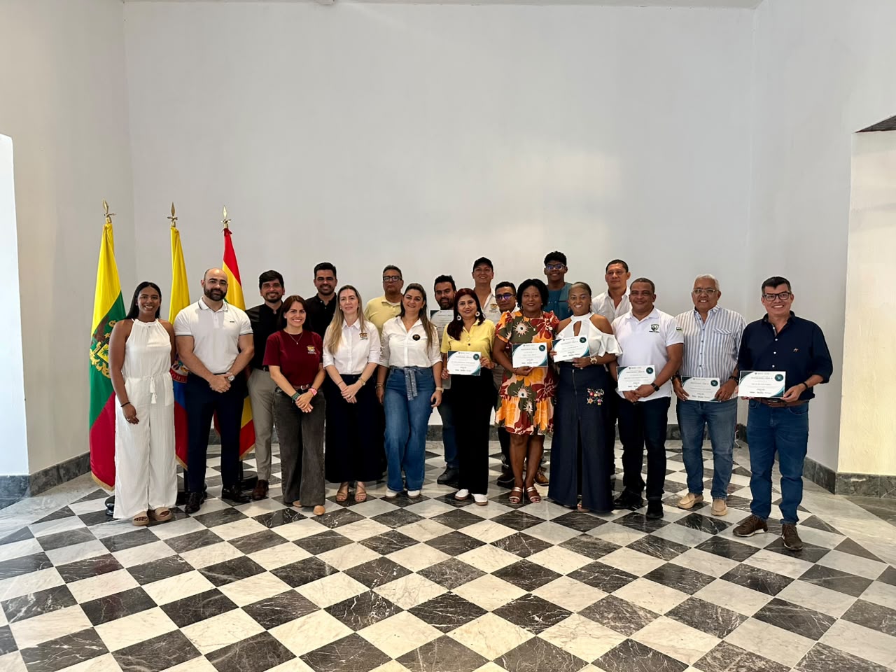 Grupo de participantes posa con diplomas junto a banderas en un salón de piso ajedrezado.