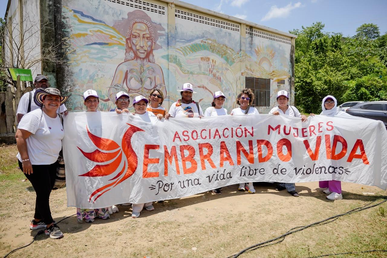 Mujeres sostienen una pancarta de la Asociación Mujeres Sembrando Vida frente a un mural.