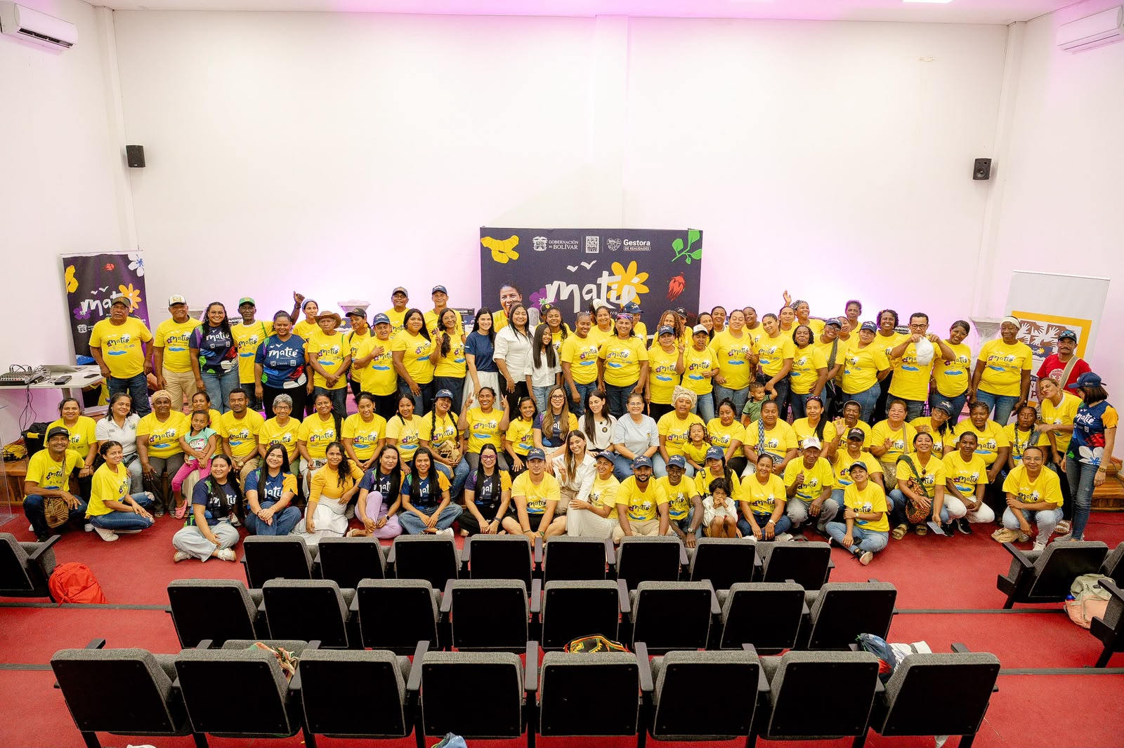Grupo numeroso de personas con camisetas amarillas del proyecto MATIE posa en un auditorio.