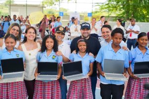 Estudiantes con uniforme escolar sostienen computadores portátiles junto a funcionarios durante la entrega.