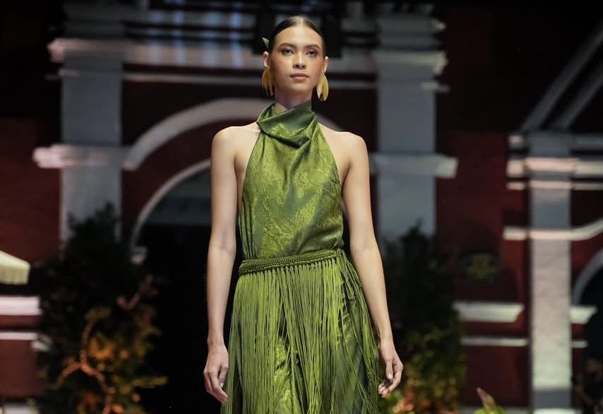 Modelo en pasarela con vestido verde de tela plisada, frente a una fachada de arcos.